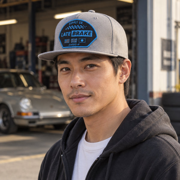Man wearing a Late Brake light grey flat brim cap with a blue LATE BRAKE patch outdoors.