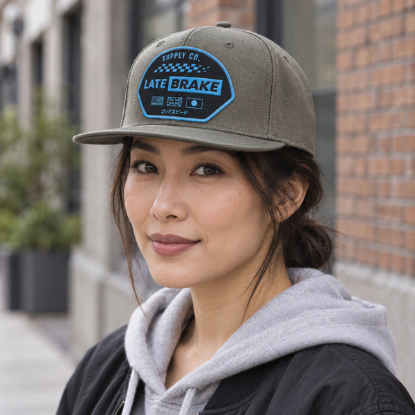 Woman wearing a Late Brake olive green flat brim cap with a blue LATE BRAKE patch outdoors.