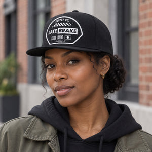 Woman wearing a Late Brake black flat brim cap with a white LATE BRAKE patch outdoors.
