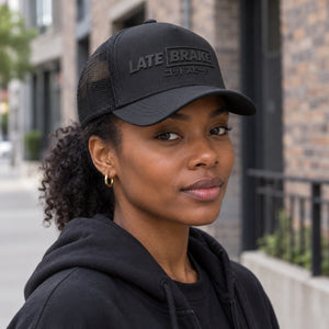 Woman wearing a Late Brake black mesh trucker hat with a tonal LATE BRAKE wordmark outdoors.