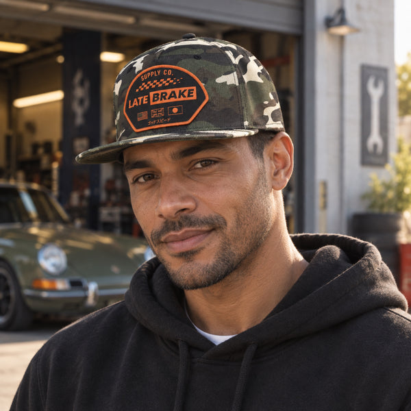 Man wearing a Late Brake woodland camo flat brim cap with an orange LATE BRAKE patch outdoors.