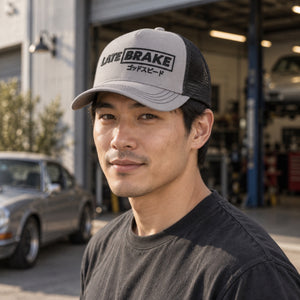 Man wearing a Late Brake grey and black mesh trucker hat with a black LATE BRAKE wordmark outdoors.